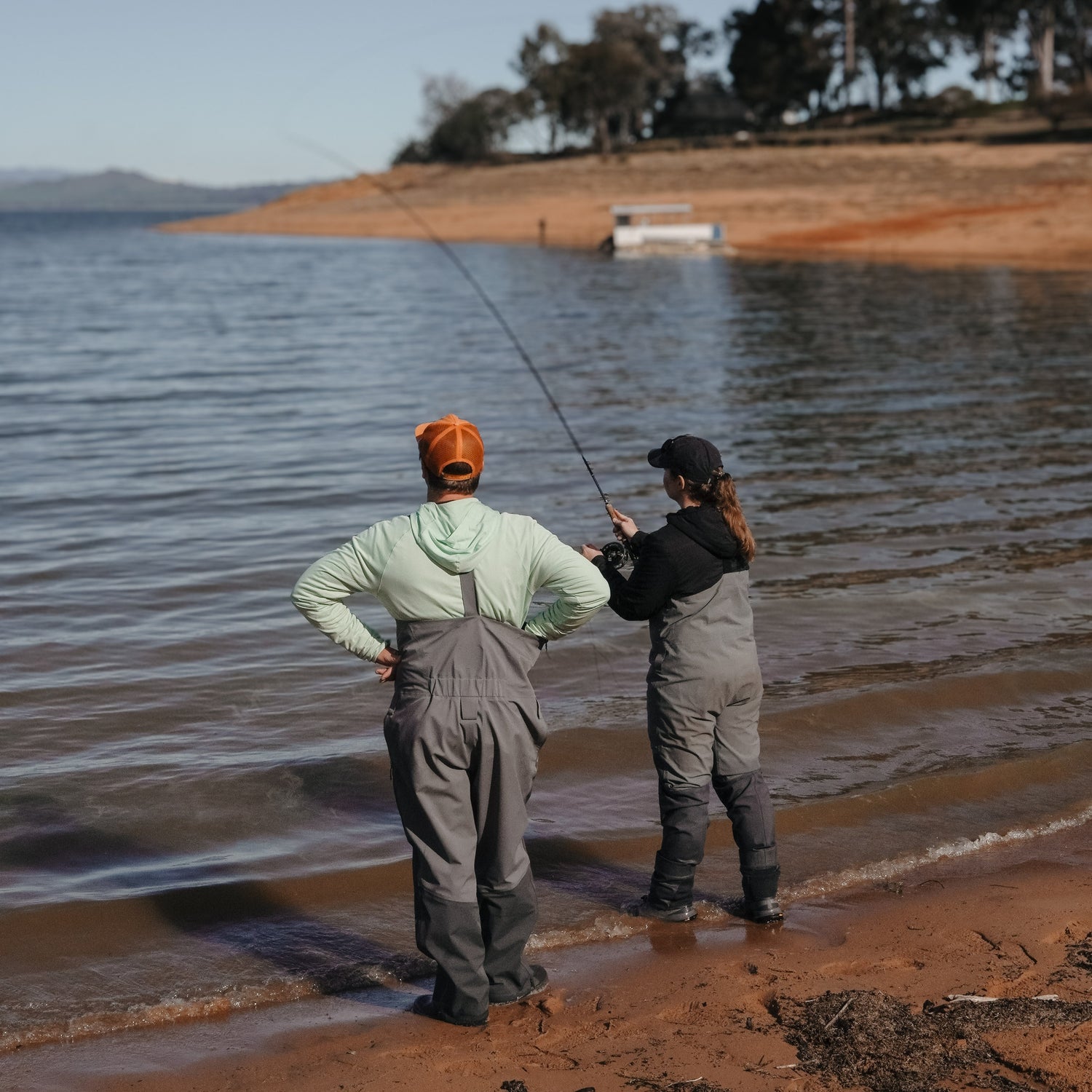 Two people fishing by a lake with trees and mountains in the background.